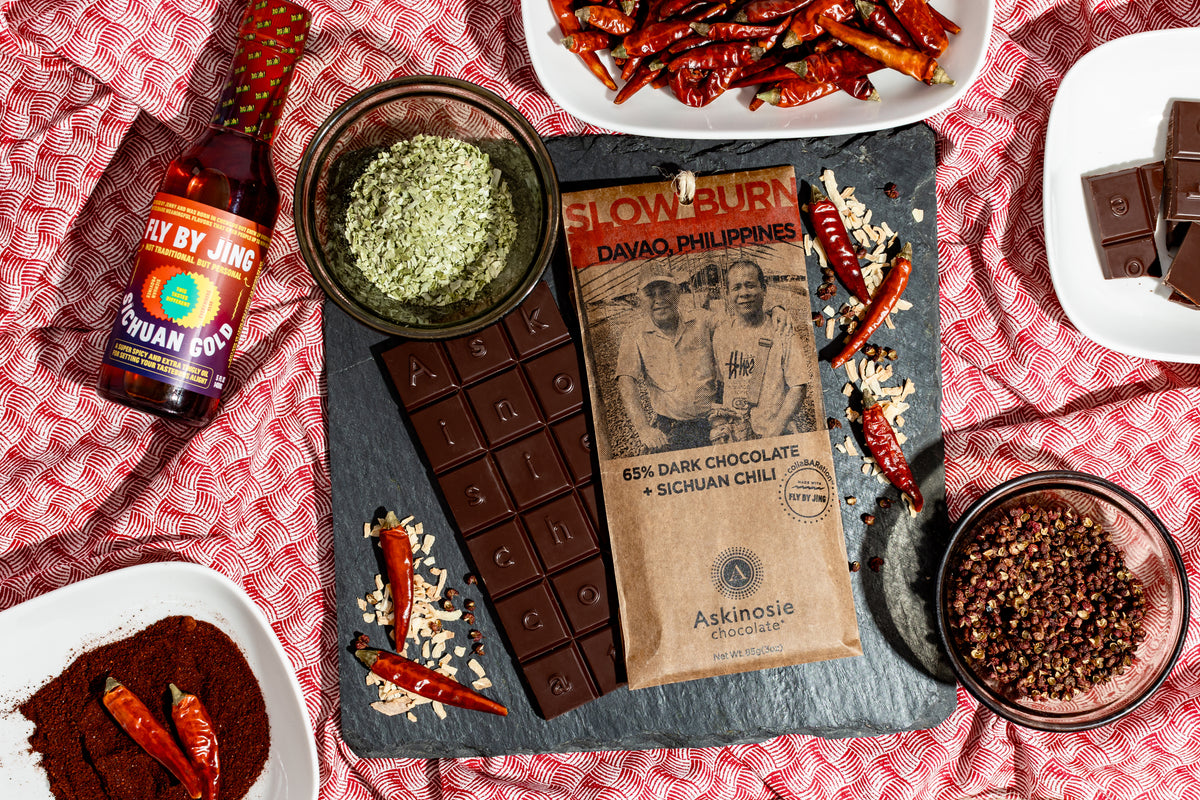 Chili peppers and various ingredients spread around the kraft brown packaging with "Slow Burn" printed in red lettering and Davao, Philippines farmers featured on the front of the Askinosie Chocolate bar wrapper.